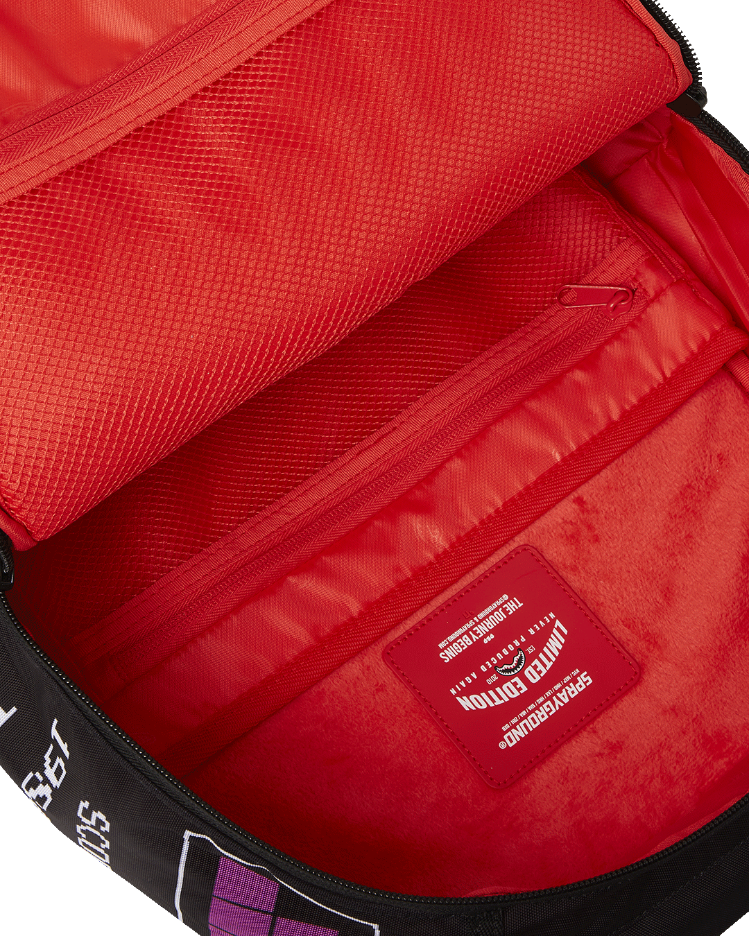 A close-up, internal view of the main compartment of a black Sprayground backpack. The interior is bright red. On the inside of the front flap, there is a large, red mesh zippered pocket. Below it, a smaller red mesh pocket is visible. On the back wall of the compartment, there is a red, fleece-lined laptop sleeve. A red, rectangular tag with a shark mouth logo reads "LIMITED EDITION" and "SPRAYGROUND" in white lettering.