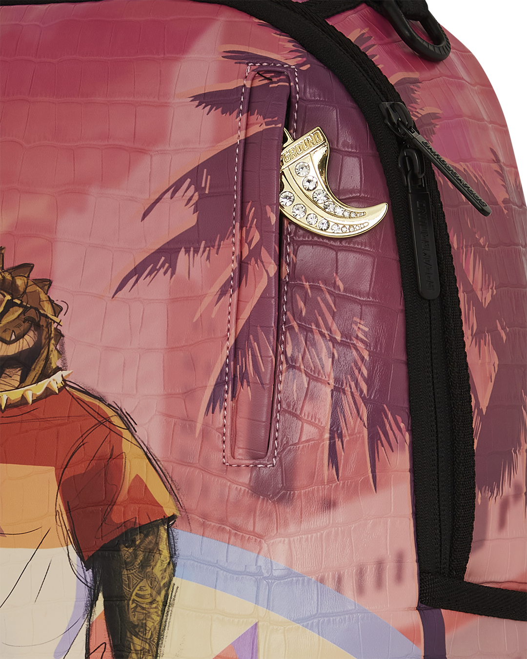 A close-up of a vibrant Sprayground backpack. The image focuses on the top right side, showing a gold charm in the shape of a tooth or tusk with embedded rhinestones, hanging from a side pocket zipper. The backpack's surface has a crocodile-skin texture and a graphic of a palm tree silhouetted against a pink and orange sunset. A black zipper with a black pull is visible on the edge.