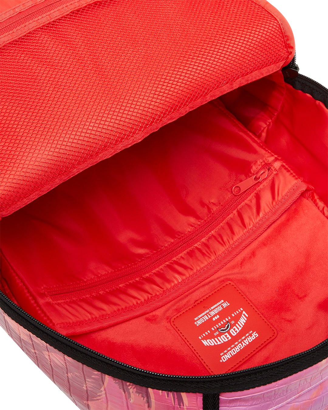 A close-up, internal view of the main compartment of a Sprayground backpack. The interior is a bright red and appears to be made of satin or a similar smooth material. On the inside of the front flap, there is a large, red mesh zippered pocket, and below it, a smaller red mesh pocket. The back wall of the compartment has a fleece-lined pocket for a laptop or tablet. A red, rectangular tag with a shark mouth logo reads "LIMITED EDITION" and "SPRAYGROUND" in white lettering. The outside edge of the backpac...