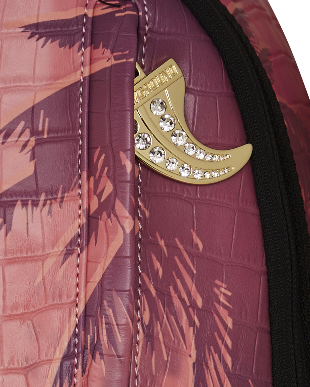 A close-up of a vibrant Sprayground backpack. The image focuses on the top right side, showing a gold charm in the shape of a tooth or tusk with embedded rhinestones, hanging from a side pocket zipper. The backpack's surface has a crocodile-skin texture and a graphic of a palm tree silhouetted against a pink and orange sunset. A black zipper with a black pull is visible on the edge.