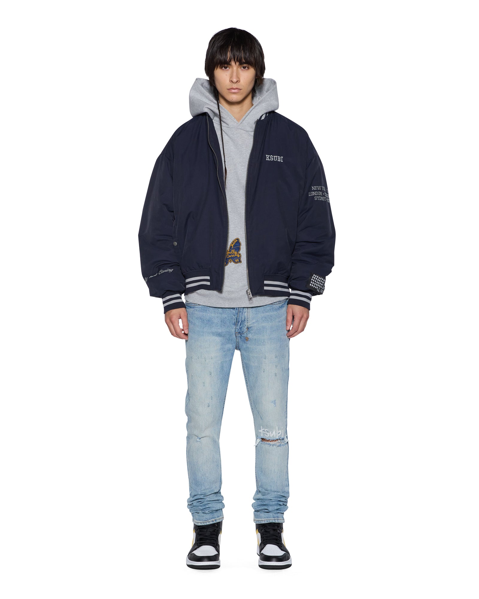 Person wearing a navy bomber jacket with gray hood, gray shirt, and light blue jeans on a white background