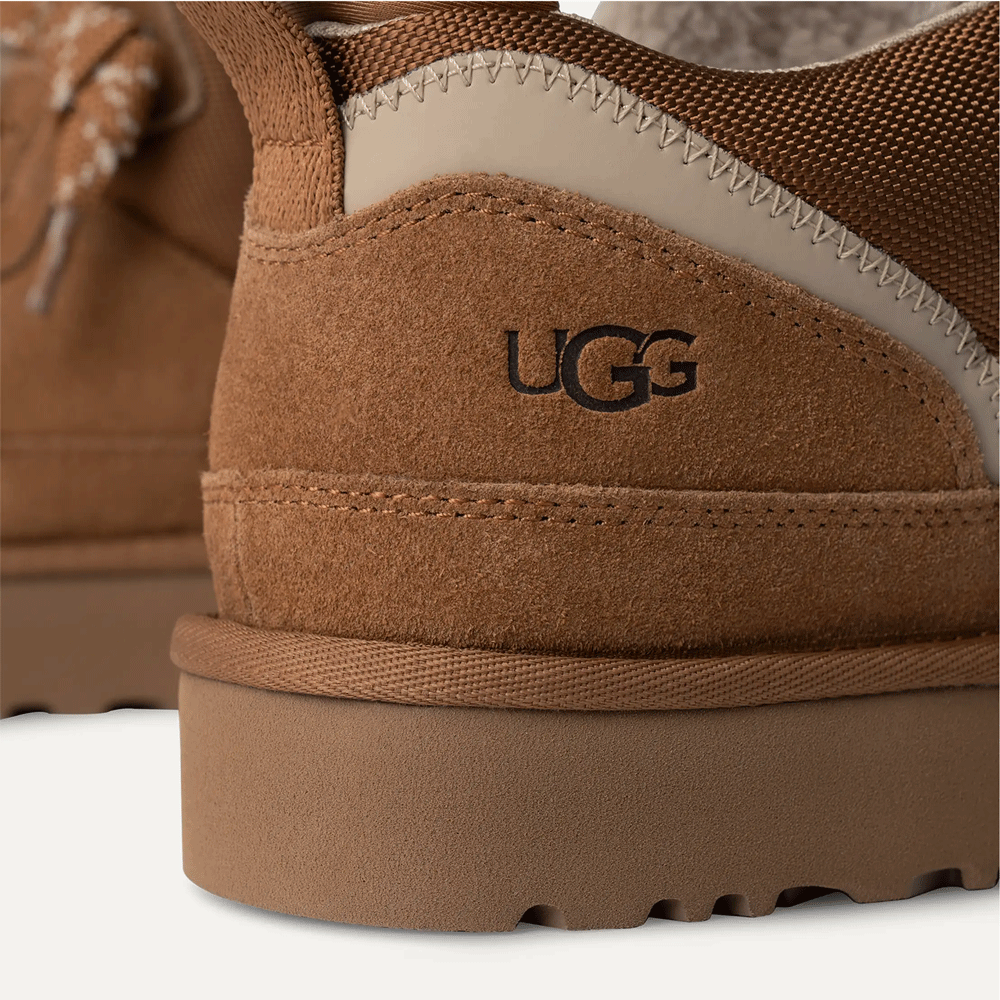 Close-up of the heel of the chestnut UGG low-top shoe, focusing on the black embossed UGG logo on the suede panel, the contrasting tan synthetic trim, and the texture of the thick platform sole.