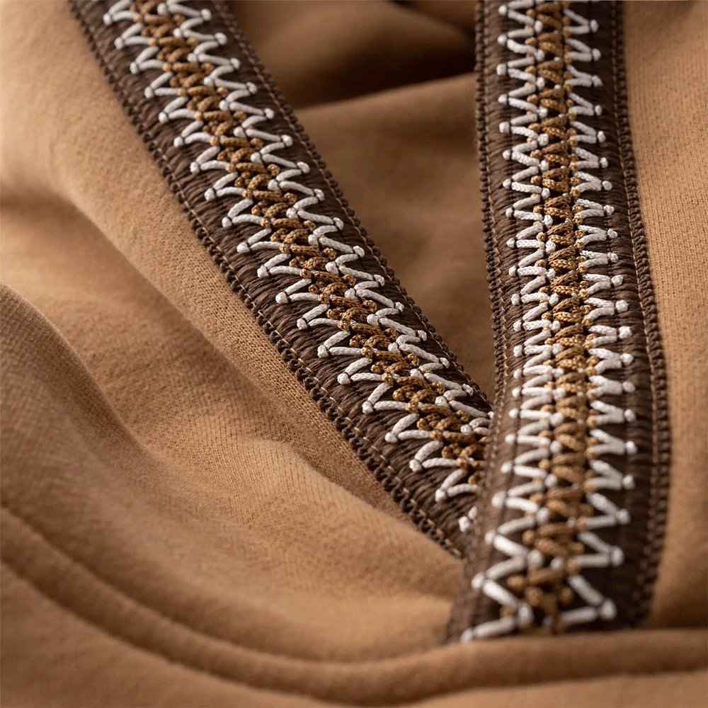 Extreme close-up detail of the UGGbraid trim on the brown Tasman fabric. The trim features a rich, three-color zig-zag pattern in tan, and white stitching, sewn onto the edge of the material.