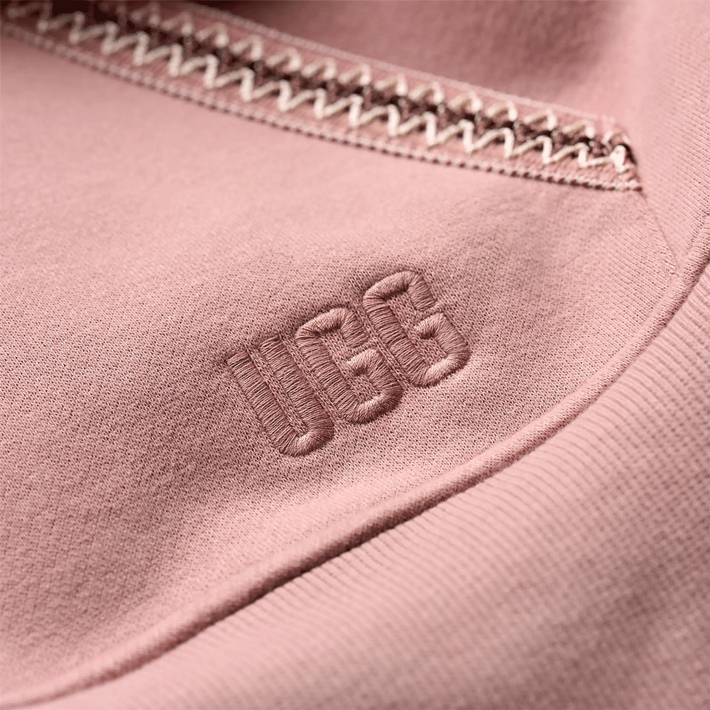 Close-up of the embroidered UGG logo and white zig-zag trim detail on the dusty rose hoodie fabric