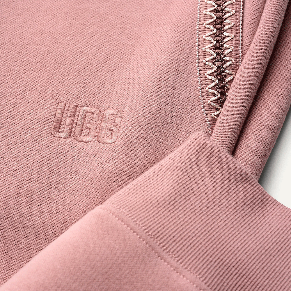 Close-up of the embroidered UGG logo on the dusty rose sweatpants, next to the ribbed cuff and braided pocket trim.