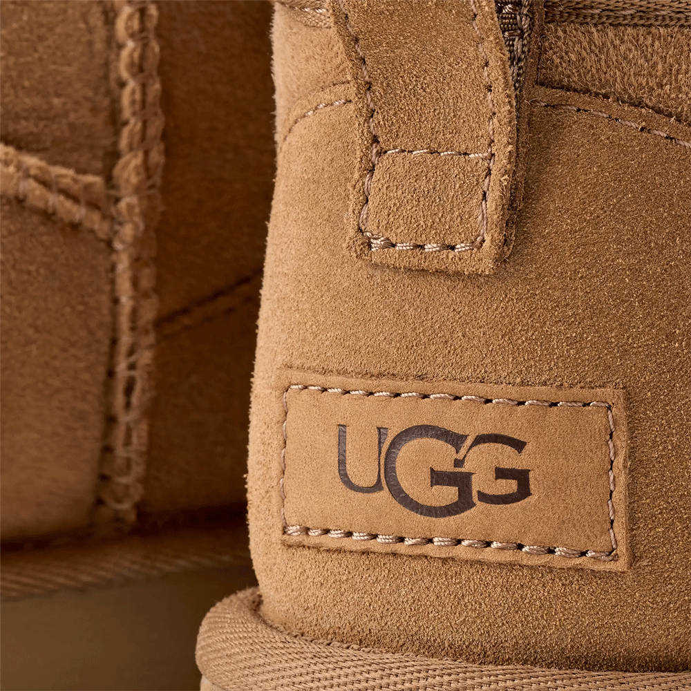 Extreme close-up of the heel of an UGG Classic Ultra Mini boot in chestnut suede. The image clearly shows the stitched rectangular UGG logo patch in a lighter brown tone and the detailed stitching of the suede heel pull tab above it.