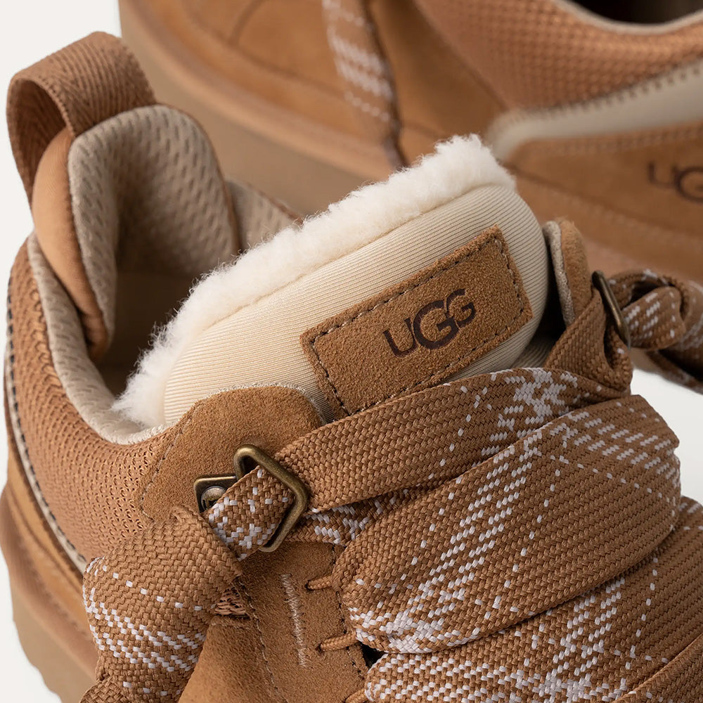 close-up view of the UGG Lowmel sneaker in chestnut, focusing on the detail of the plaid-patterned lace/strap, the exposed UGGplush wool lining, and the UGG logo patch on the oversized tongue.