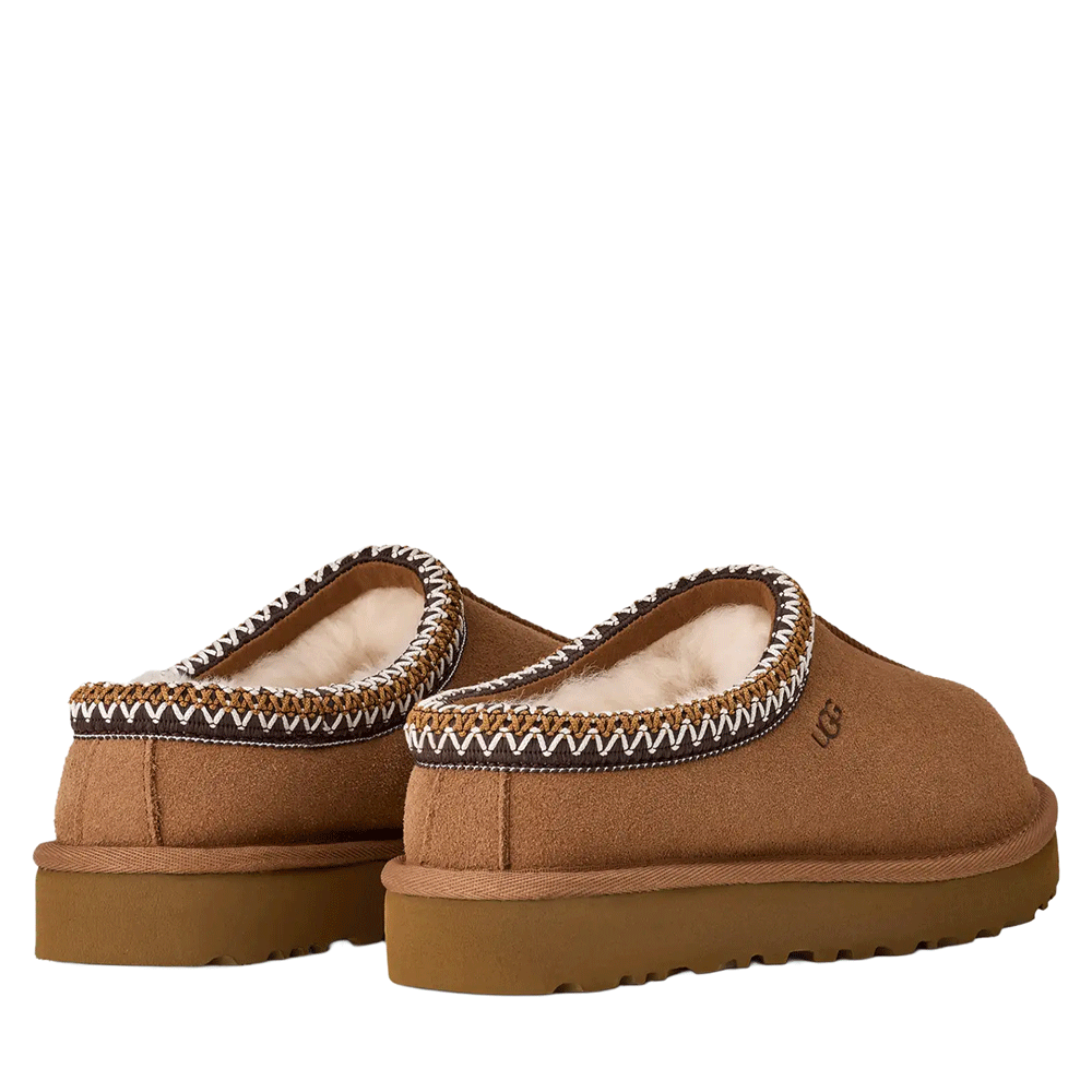 A pair of UGG Tasman II slippers in brown suede, shown from a rear three-quarter view. The image highlights the cozy, natural sheepskin lining visible inside the shoe and the signature UGGbraid trim around the low collar.