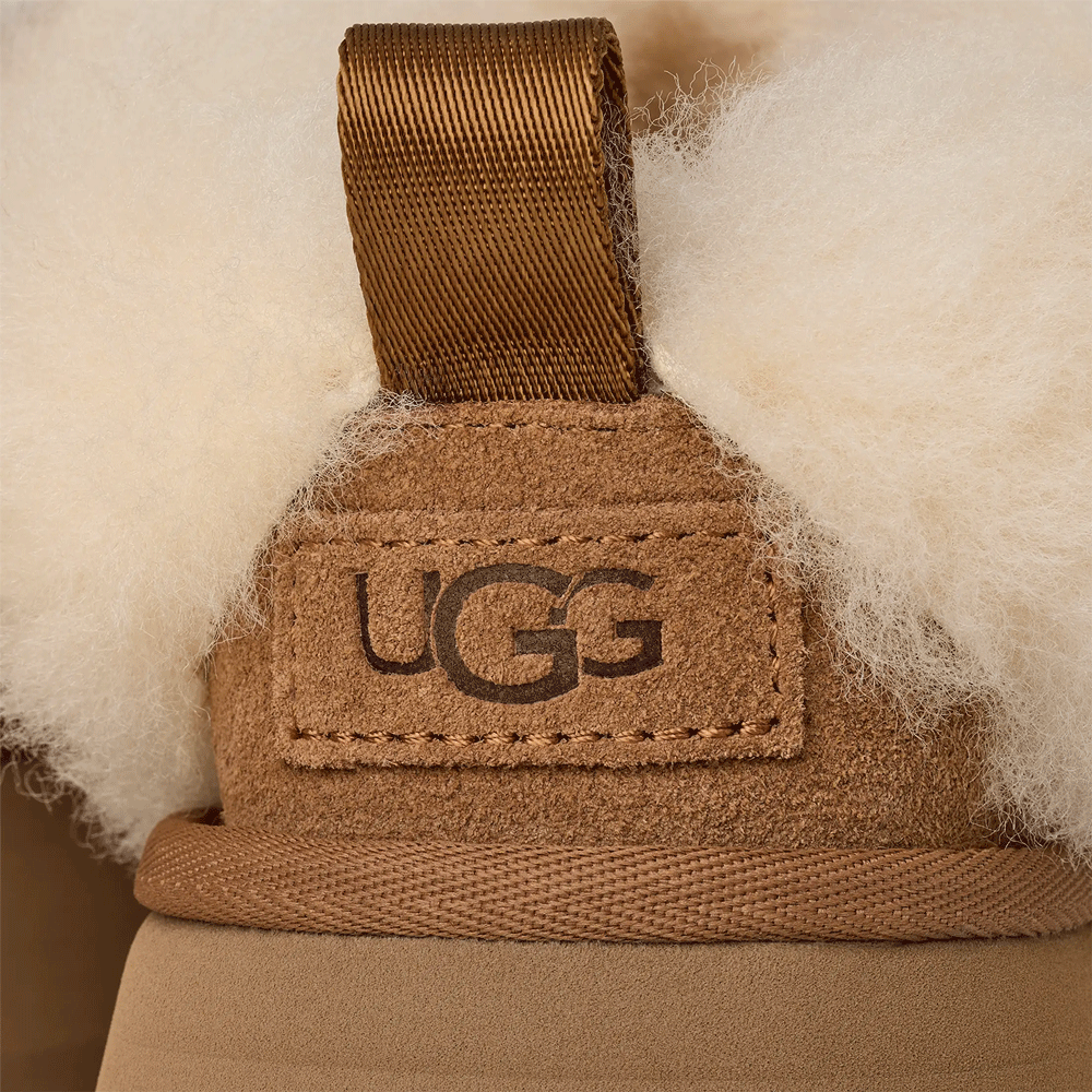 Extreme close-up on the back heel of the Chestnut UGG Tazzelle slipper. The image highlights the embossed UGG logo on the suede heel label, the fabric pull tab, and the surrounding fluffy sheepskin collar.