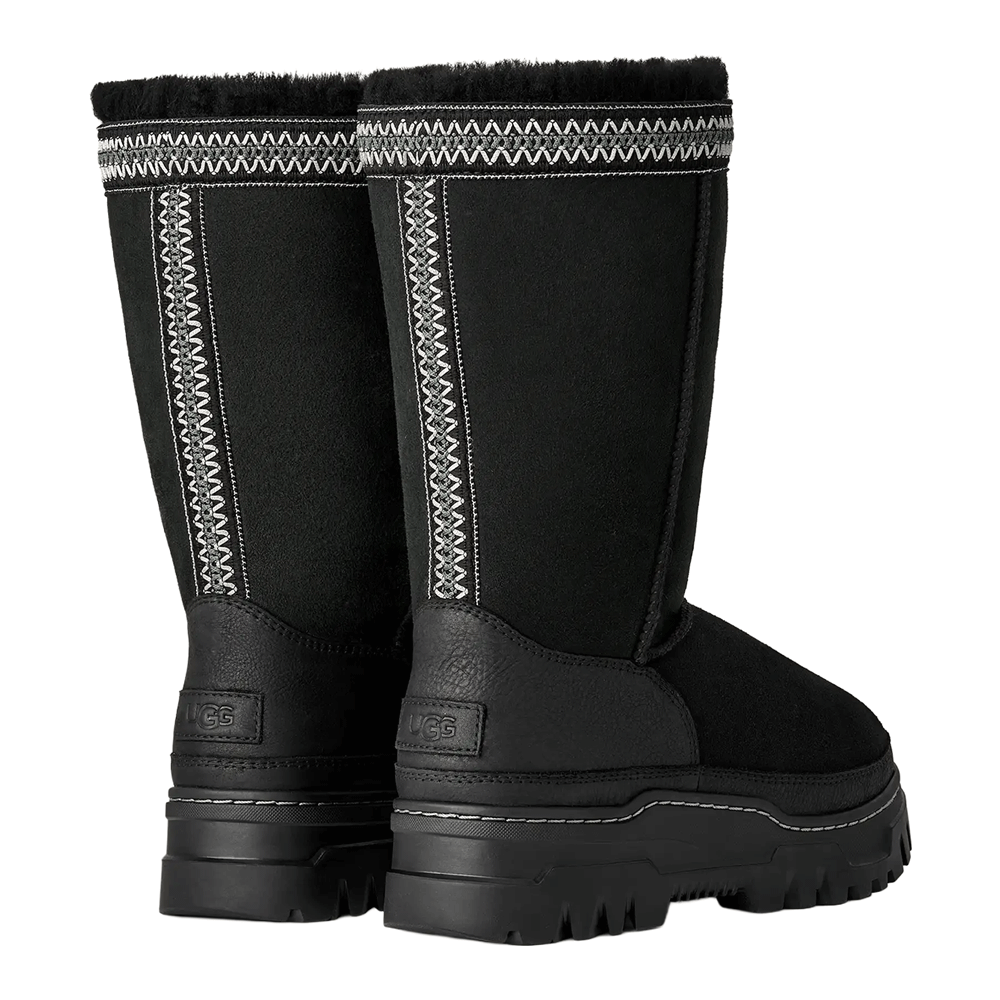 Close-up of the black UGG low-top shoe tongue and laces, focusing on the black UGG logo patch, the metal lace hardware, and the thick, black and white speckled mountaineering-style laces.