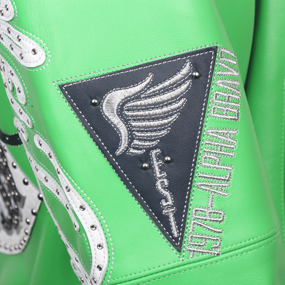 Close-up of a triangular patch on the sleeve of a green Pelle Pelle jacket. The patch features a silver embroidered wing and sword on a black leather base with silver studs. Vertical silver embroidery next to the patch reads "1978-ALPHA BRAVO," further highlighting the brand's founding year and military-inspired theme.