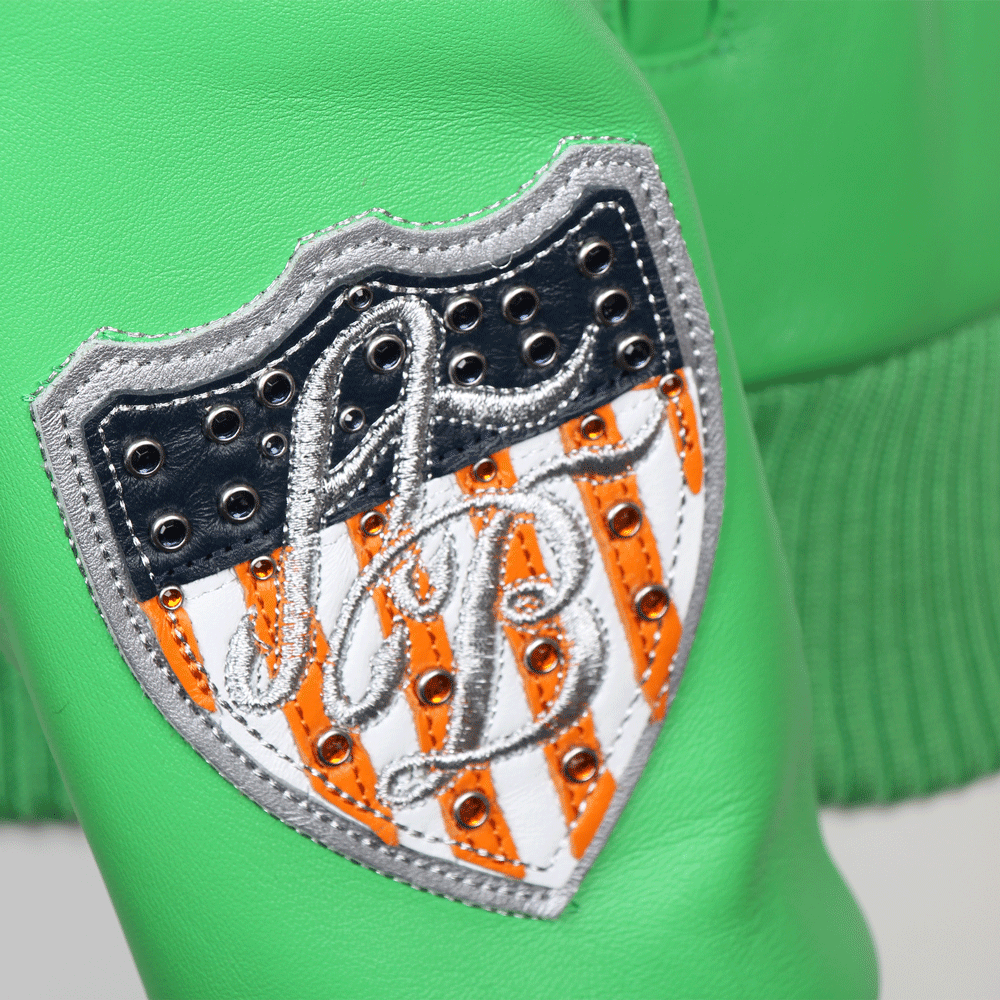 A detailed close-up of a shield-shaped leather patch on a green Pelle Pelle jacket. The patch features a silver embroidered "PB" monogram over orange and white vertical stripes, encased in a black leather border with metallic silver studs and orange crystals.
