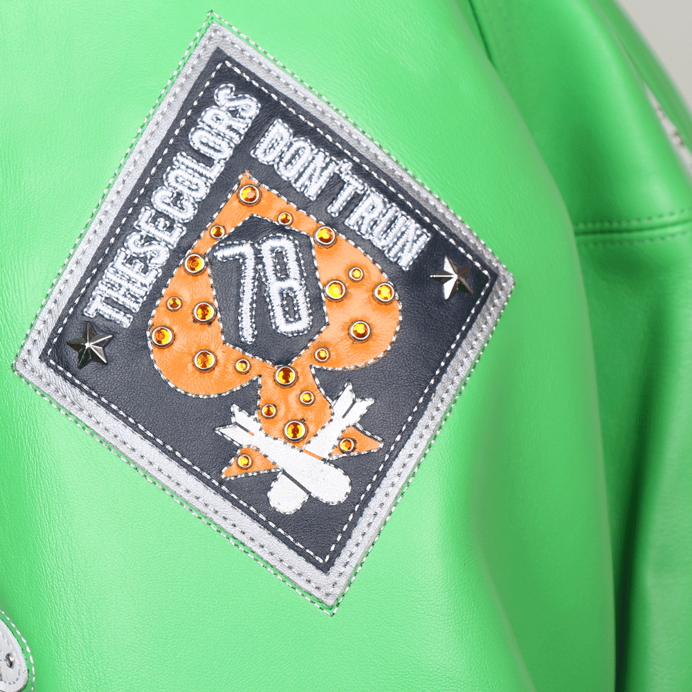 A close-up of a diamond-shaped leather patch on the sleeve of a green Pelle Pelle jacket. The patch features the slogan "THESE COLORS DON'T RUN" in silver embroidery surrounding an orange spade with the number "78" in the center. The design is accented with orange crystals and silver metallic studs.