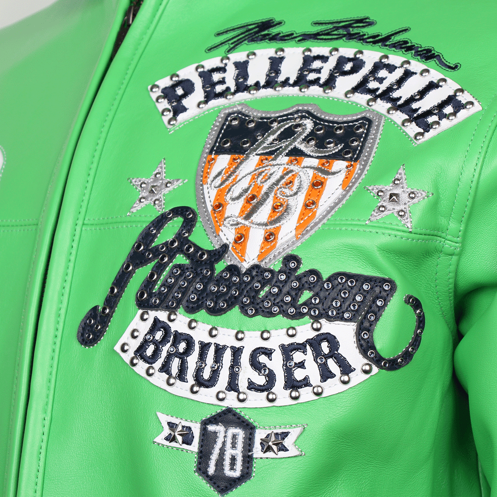 A detailed close-up of the chest embroidery on a green Pelle Pelle American Bruiser leather jacket. The image shows a multi-layered "American Bruiser" patch with silver studs, a shield featuring orange and white stripes, and the "Pelle Pelle" brand name arched above in a studded script.