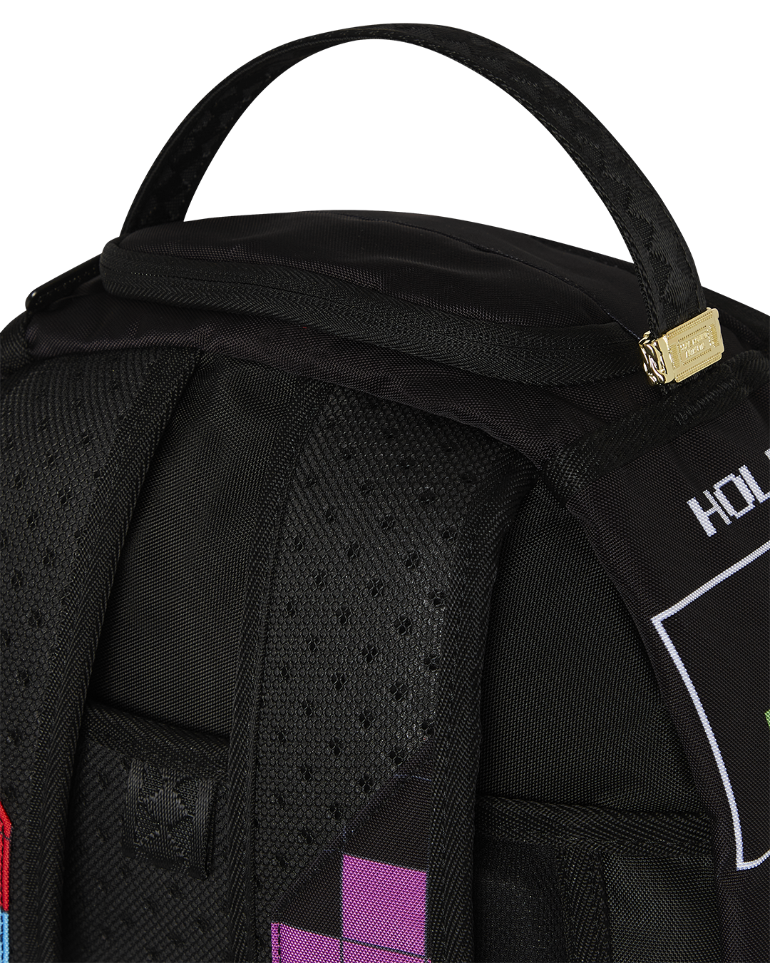 A close-up of the top portion of a black Sprayground backpack. The image shows the padded, black top handle with a textured pattern. A gold-toned metal tag with the Sprayground logo is attached to the handle. The top of the backpack features a zippered pocket. Below the handle, the back panel is black and padded with a dotted texture, and a black shoulder strap is visible. A purple and a pink Tetris block can be seen on the bottom edge of the image.
