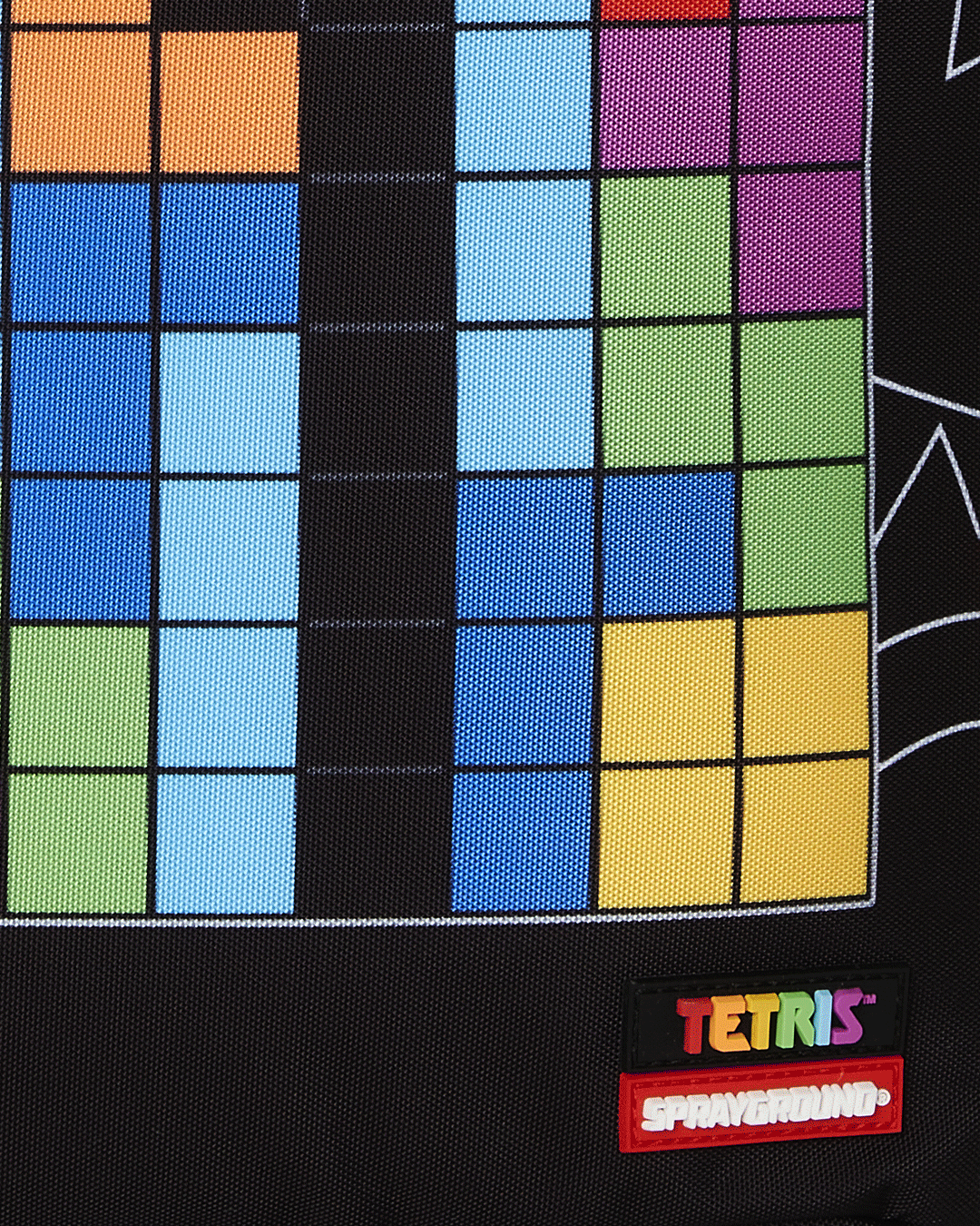 A close-up of the bottom of a black Sprayground backpack, showing a vibrant, pixelated Tetris grid design. The grid is filled with various colored blocks, including blue, orange, light blue, green, and yellow. Below the grid, a red and black tag is sewn onto the backpack. The tag has the Tetris logo in colorful letters and the Sprayground logo in white lettering on a red background.
