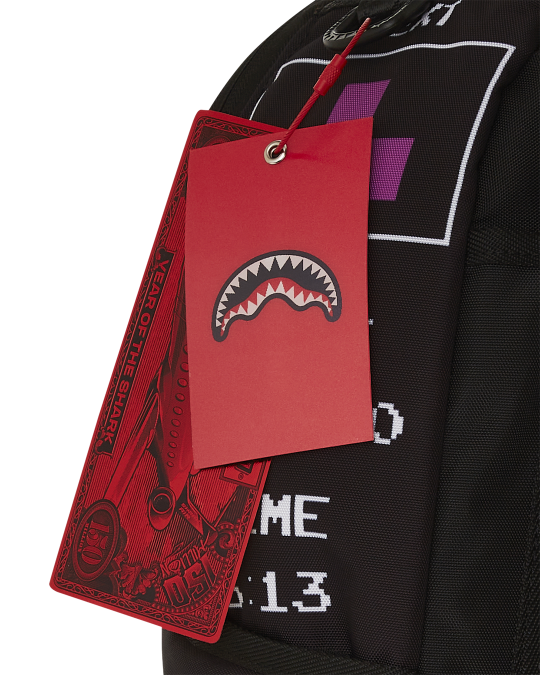 A close-up of two red tags hanging from a black Sprayground backpack. The front tag is a solid red rectangle with the brand's signature shark mouth logo in white. The back tag is long and has a banknote-like design with the words "YEAR OF THE SHARK" and a large "150" on it. Part of the backpack is visible with a white graphic that says "TIME 6:13" and a purple Tetris block in a white box.
