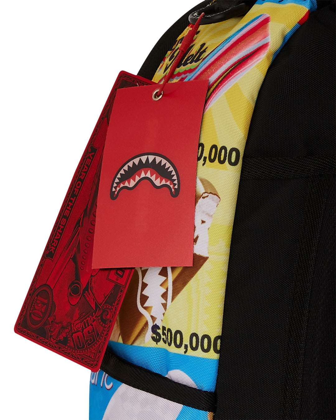 A close-up of two red tags hanging from a colorful Sprayground backpack. The front tag is a solid red rectangle with the brand's signature shark mouth logo in white. The back tag is long and has a banknote-like design with the words "YEAR OF THE SHARK" and a large "150" on it. Part of the backpack is visible with a graphic that includes a brown zipper pull, a cartoon ice cream wrapper with a price of $500,000, and a black shoulder strap.