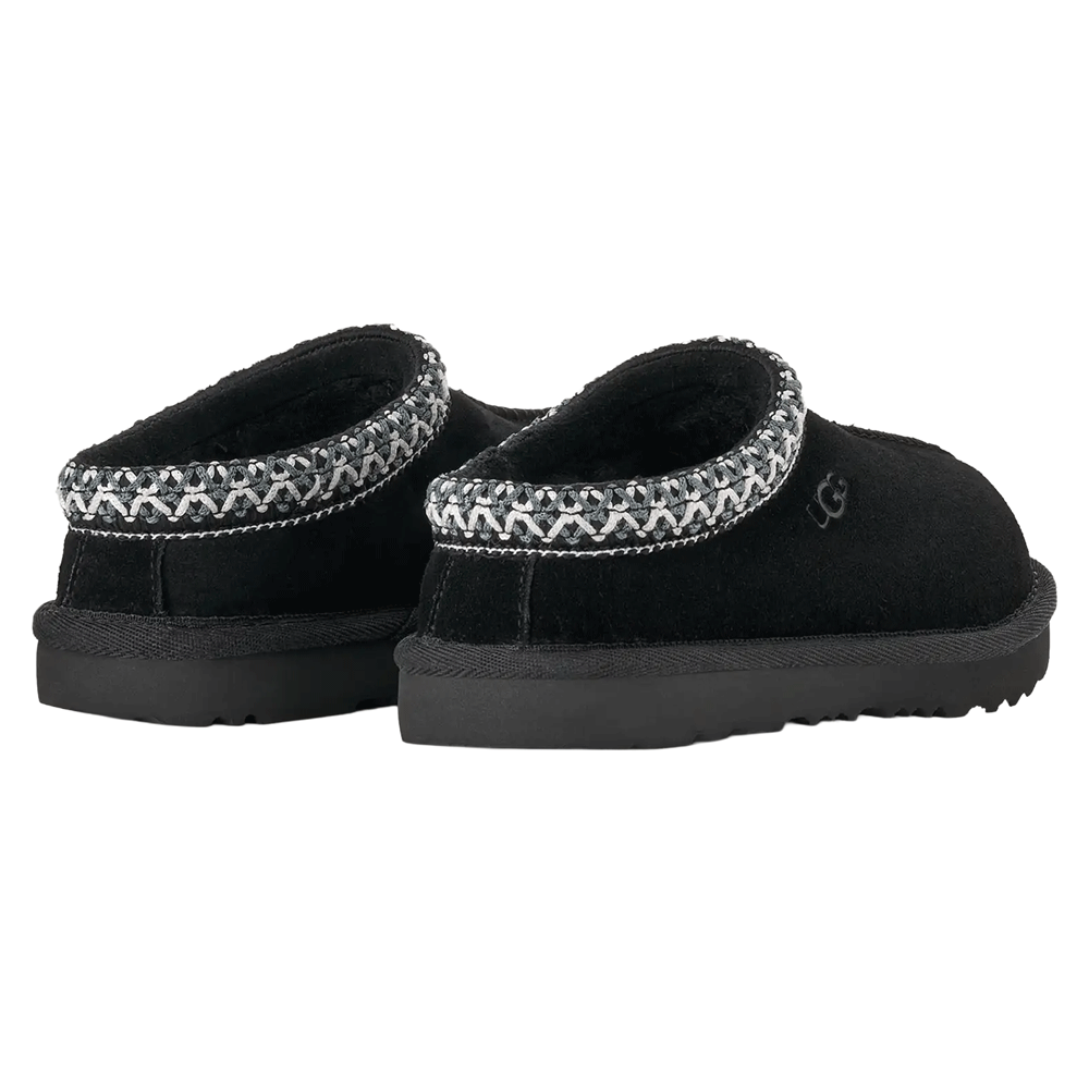 Rear angle view of a pair of black Kids' UGG Tasman slippers, showing the plush UGGplush lining visible at the opening, the black suede heel, and the full wrap-around grey and white UGGbraid detail.