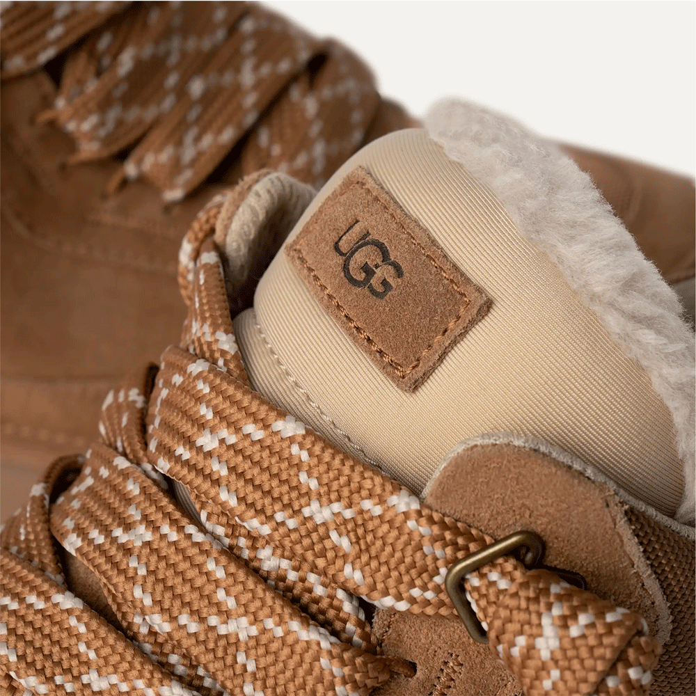 Extreme close-up of the top of the chestnut UGG low-top shoe, focusing on the UGG logo patch on the tongue, the thick, patterned tan and white mountaineering-style laces, and the plush white interior lining.