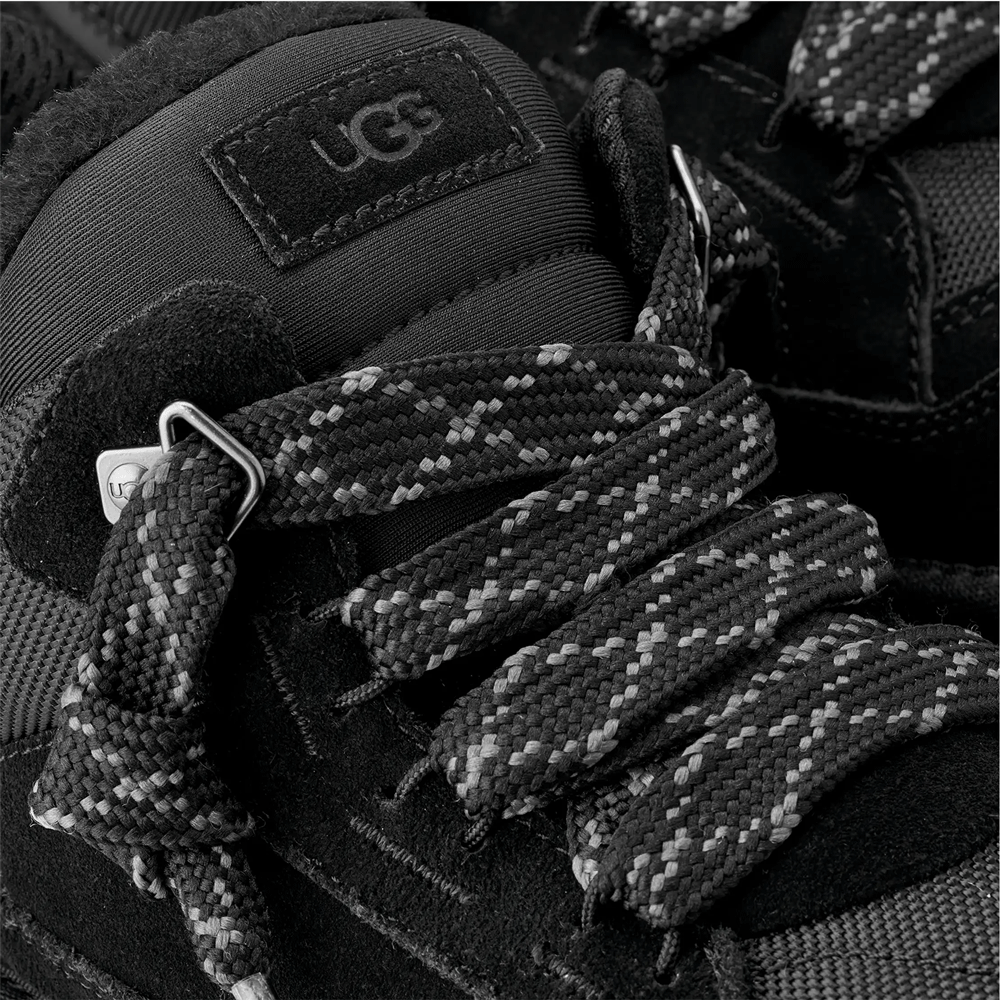 Extreme close-up of the top of the black UGG low-top shoe, focusing on the black UGG logo patch on the tongue and the thick, patterned black and white speckled mountaineering-style laces.
