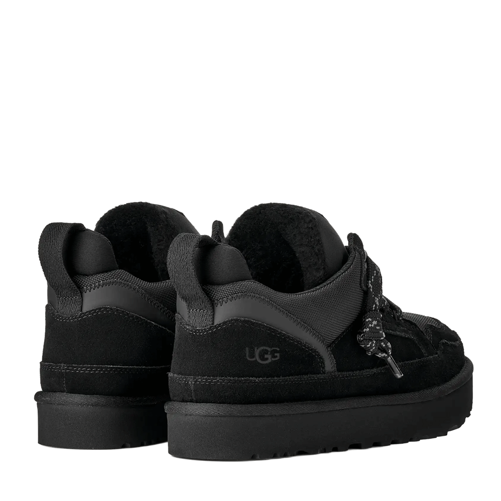 Rear view of a pair of low-top UGG shoes in black suede and textile, highlighting the black plush interior lining, the embossed black UGG logo on the heel, and the black pull tabs at the back collar.