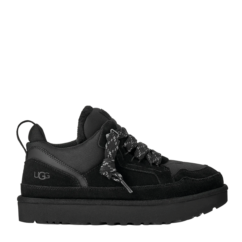 Side view of a low-top UGG shoe in all black suede and textile, featuring thick speckled black and white laces, a black plush lining visible at the collar, and a substantial black platform sole with a lugged tread.