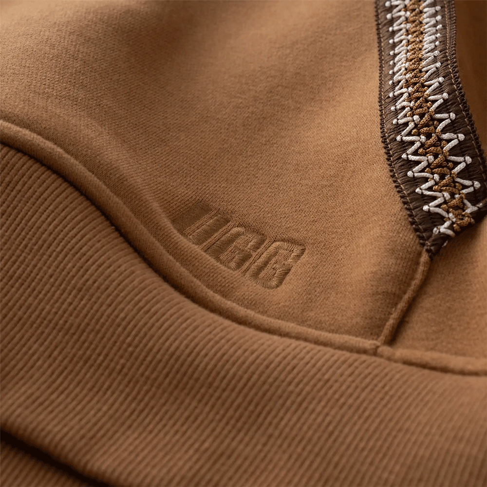 Close-up detail shot of the brown UGG Tasman fabric, showing the textured ribbed hem or cuff, the tonal 'UGG' graphic font satin stitch embroidery, and the contrasting brown and white zig-zag UGGbraid trim.