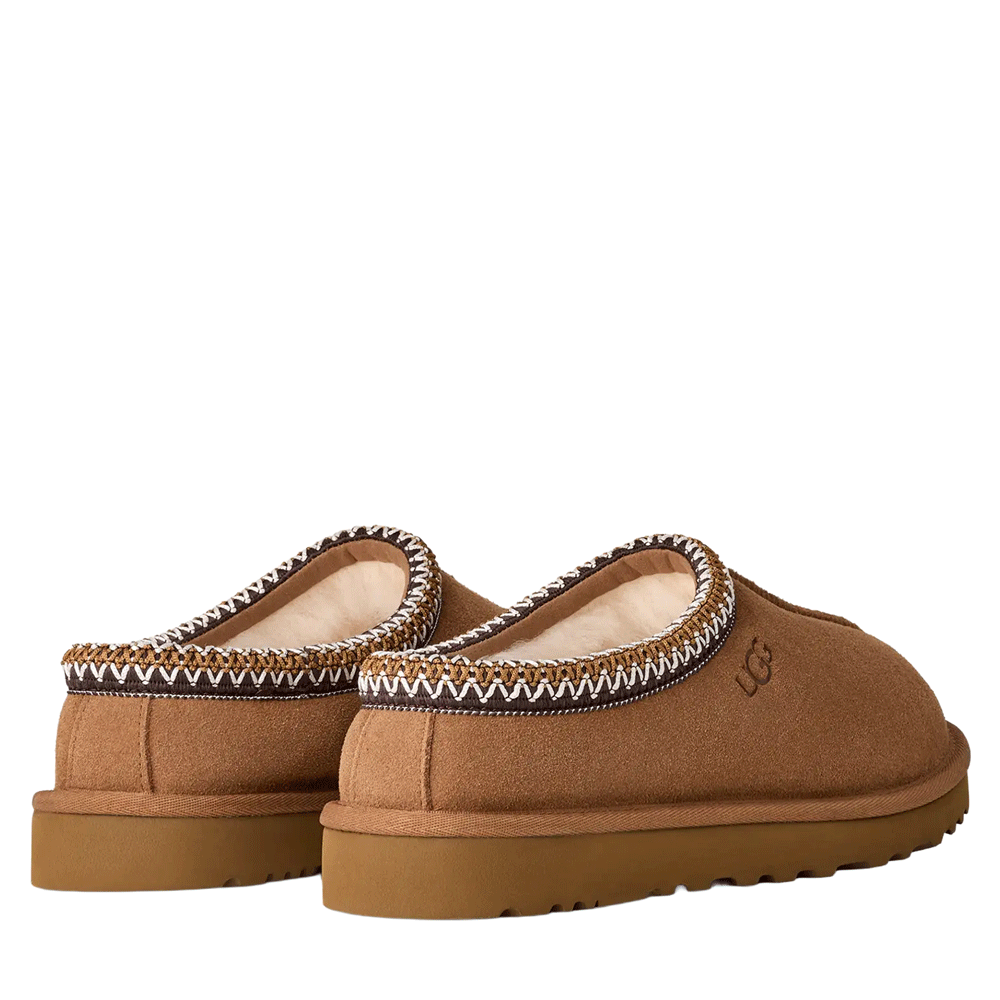Rear angle view of a pair of chestnut UGG Men's Tasman slippers, showcasing the soft suede heels, the plush, light-colored sheepskin lining visible at the collar, and the signature multi-colored UGGbraid detail.