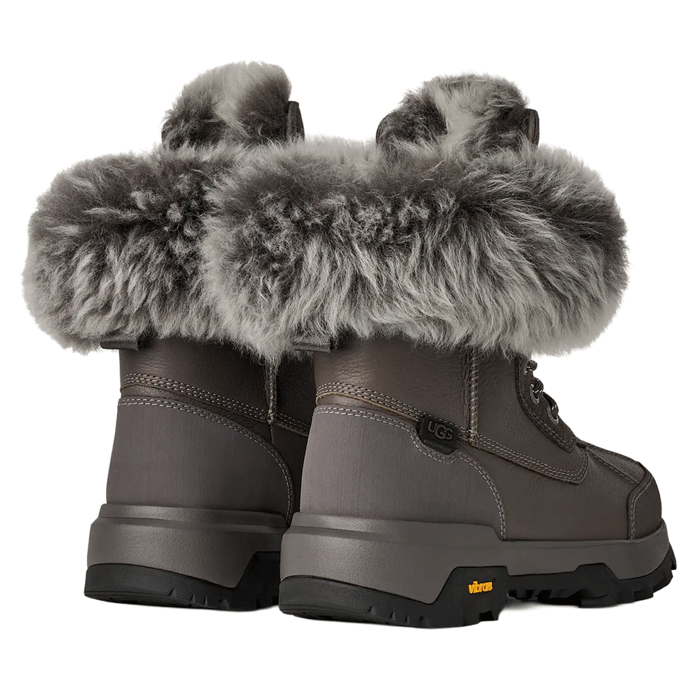 Rear view of a pair of grey UGG winter boots, emphasizing the wide, fluffy grey fur cuff, the smooth leather back panel, and the thick, lugged Vibram sole.