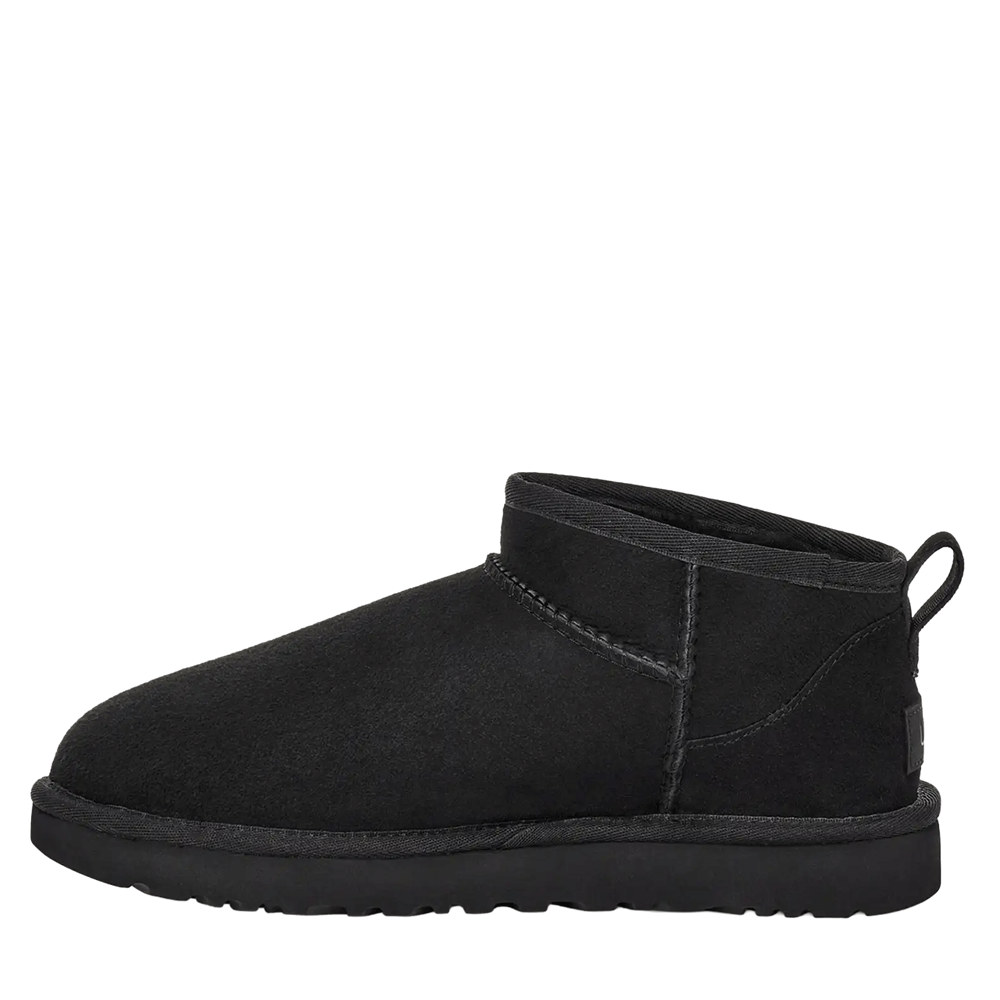 Side view of one UGG Women's Classic Ultra Mini boot in black. The image clearly shows the flat, minimalist design of the short suede shaft, the black rubber outsole, and the heel pull loop extending from the back.