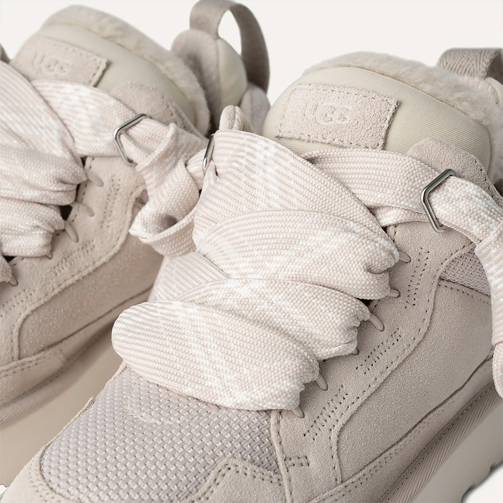 A close-up view of the front of the UGG Lowmel sneaker, highlighting the texture of the light-colored mesh vamp, the suede overlays, and the thick, two-toned plaid-patterned laces cinched loosely by the metallic eyelets.