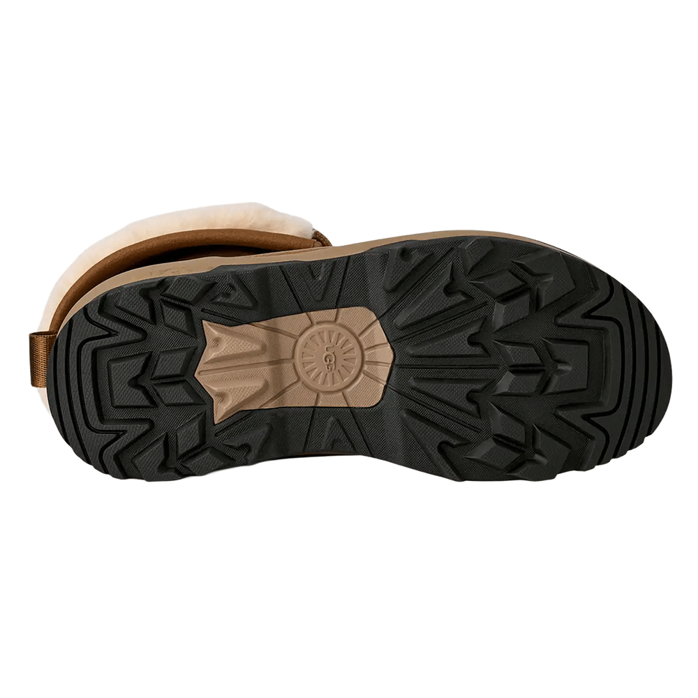 Bottom view of the UGG boot's outsole, showing the rugged, high-traction black rubber tread pattern and the tan mid-foot section featuring an embossed UGG logo.