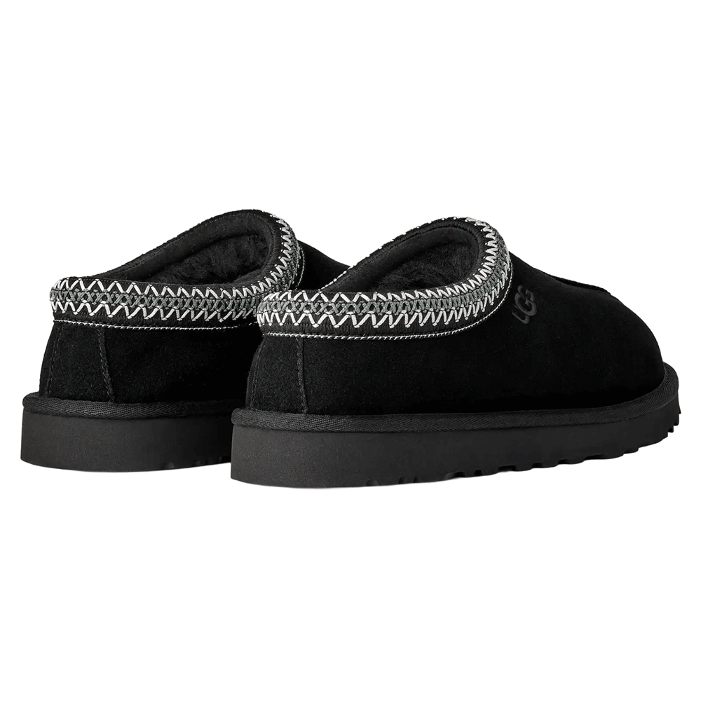 Rear view of a pair of black UGG Tasman II slippers. The image shows the heel, the dark sheepskin lining visible around the collar, and the signature gray and white UGGbraid trim. Both slippers rest on the thick, black Sugarcane EVA outsole.