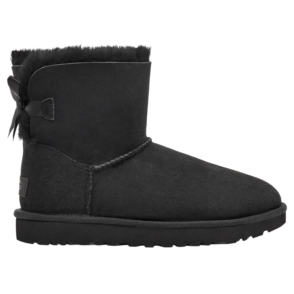 Side view of the Black UGG Mini Bailey Bow II boot, featuring a short black suede shaft with black sheepskin lining peeking out at the cuff, a fixed black satin bow along the back shaft, and a durable black Sugarcane EVA outsole.
