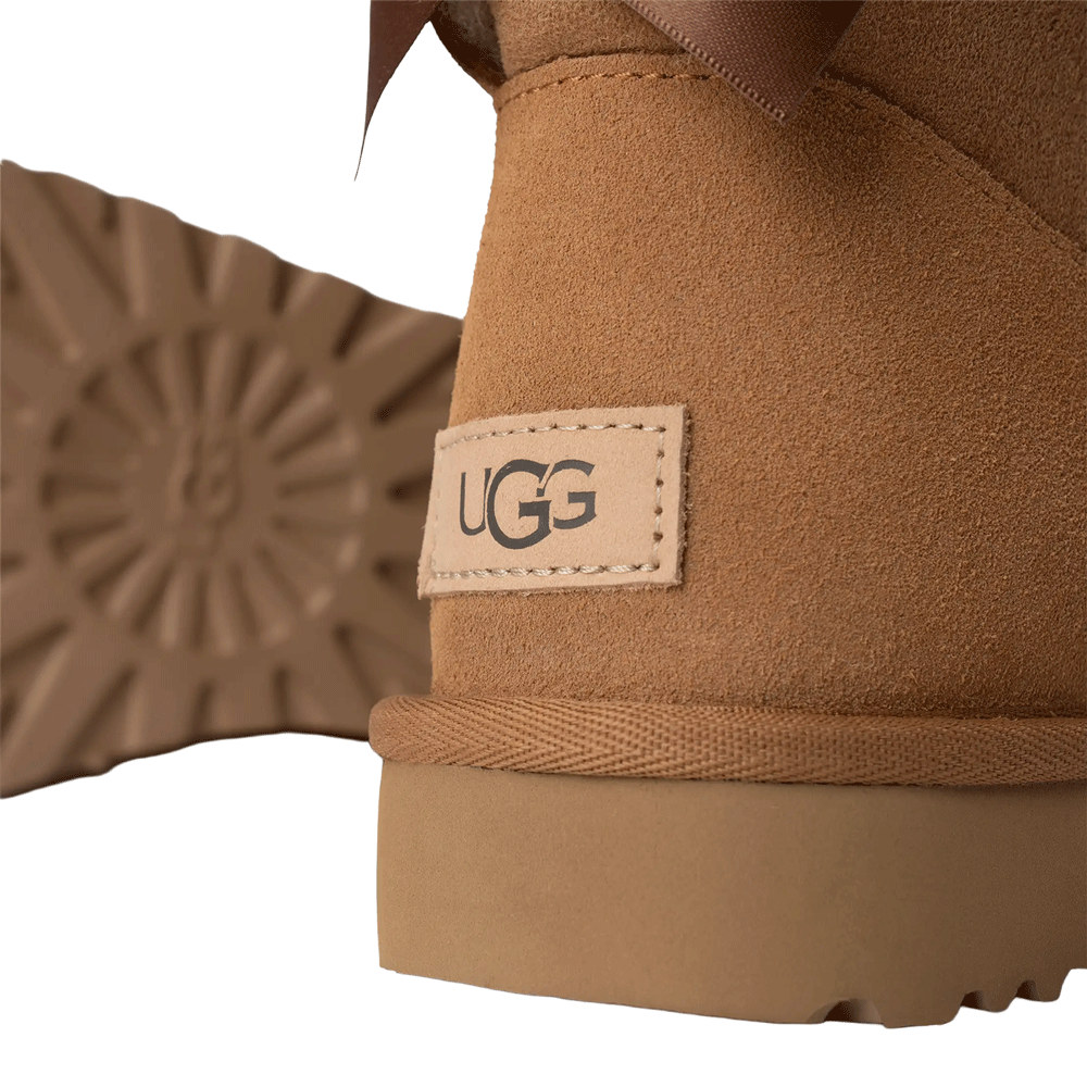 Close-up shot of the UGG logo label on the chestnut suede heel of the Mini Bailey Bow II boot, with the patterned outsole visible in the background.