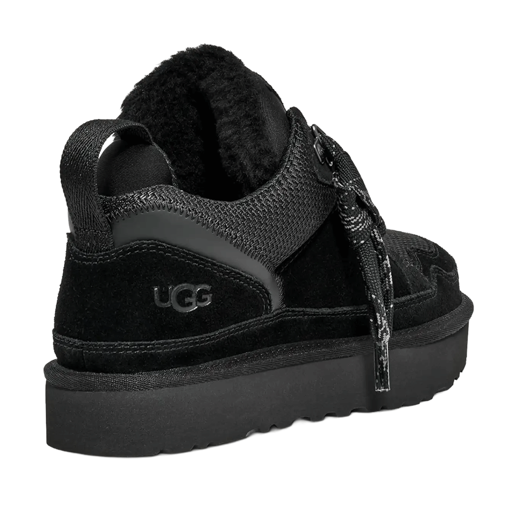 Angled rear view of the black UGG Lowmel sneaker. The shoe highlights the black suede heel counter with an embossed UGG logo, the padded collar with visible dark lining, and the thick, black Sugarcane EVA outsole. A webbing pull tab and a loose, patterned lace are also visible.