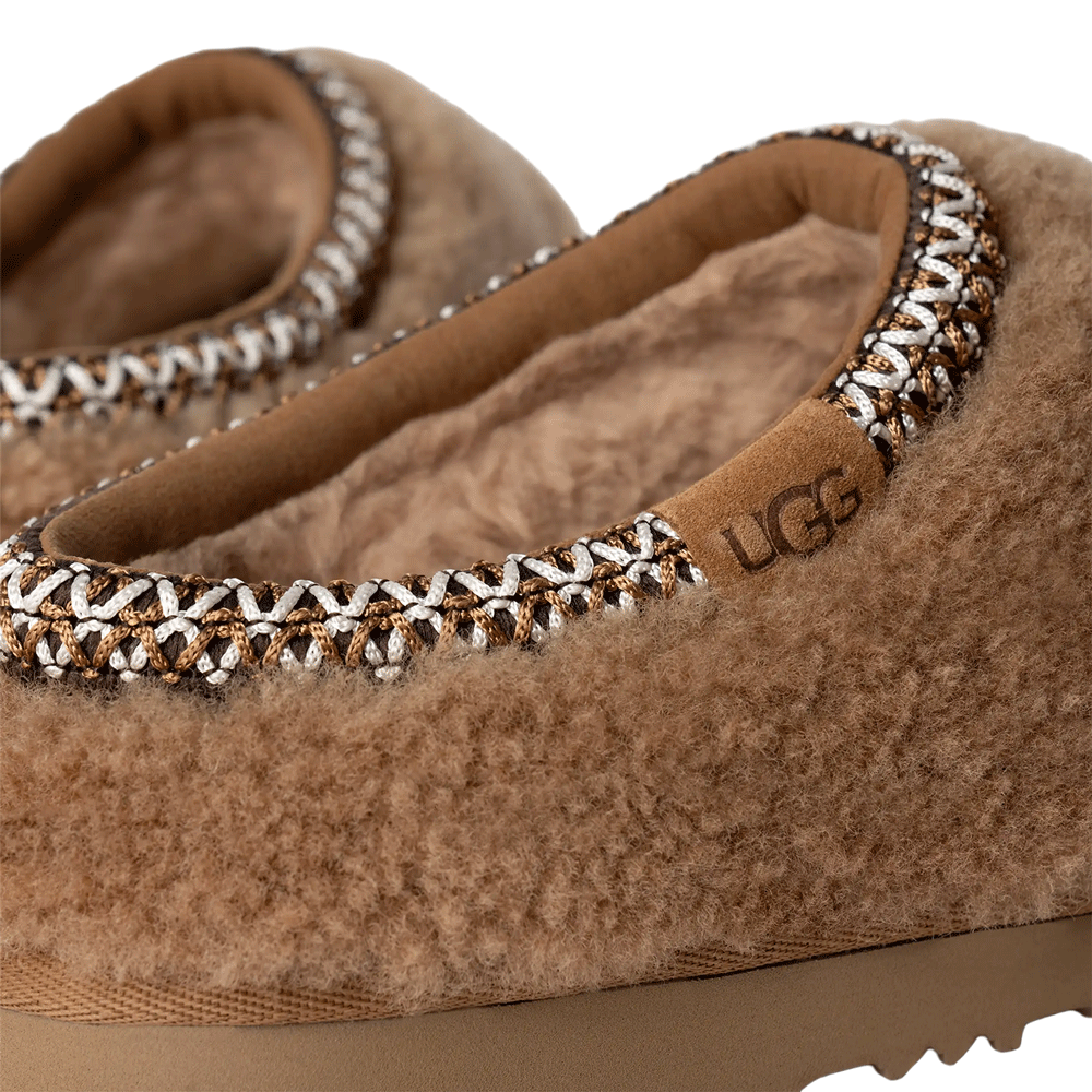 Extreme close-up of the Chestnut Tasman Maxi Curly slipper, focusing on the fluffy curly sheepskin texture, the brown suede UGG logo label, and the intricate, multi-colored UGGbraid trim around the collar.