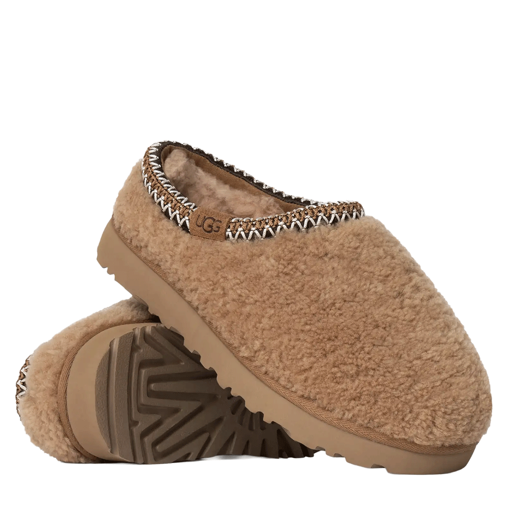 A pair of Chestnut Tasman Maxi Curly UGG slippers with fluffy sheepskin uppers, stacked on top of each other to show the woven UGGbraid trim and the lugged Sugarcane EVA outsole.