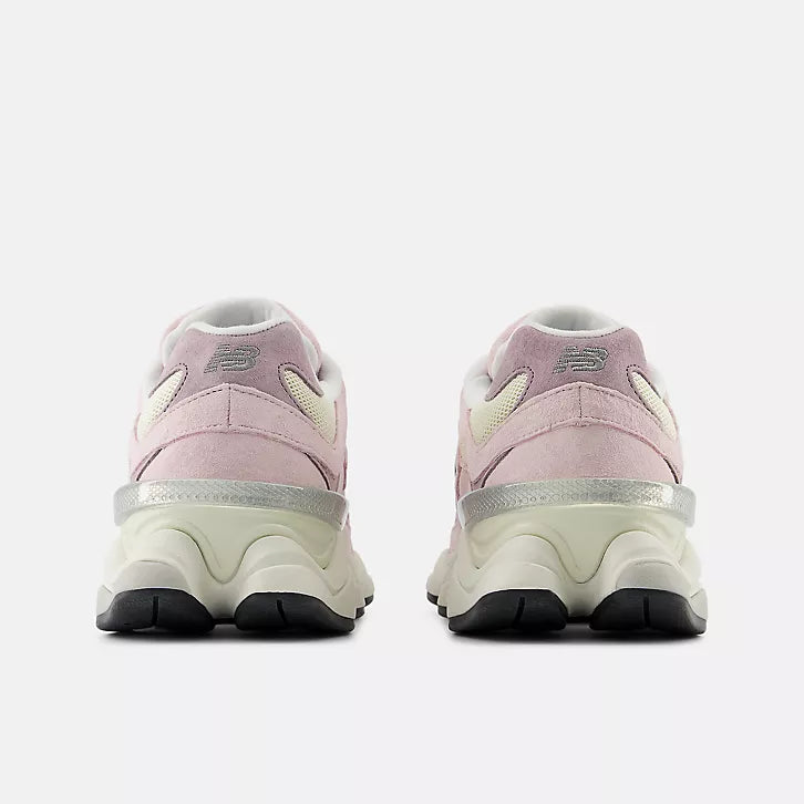 Rear view of a pair of New Balance 9060 unisex sneakers in pink and cream. The image highlights the distinctive split-pod wavy midsole in cream, the translucent metallic silver heel clip, and the embroidered grey New Balance logo on the light pink suede heel tab.