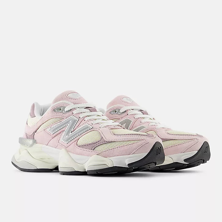 A three-quarter front view of a pair of New Balance 9060 unisex sneakers in pink and cream. The shoes feature a chunky, sculpted cream midsole with black tread, layered light pink suede and cream mesh uppers, white laces, and the signature silver "N" logo on the side.