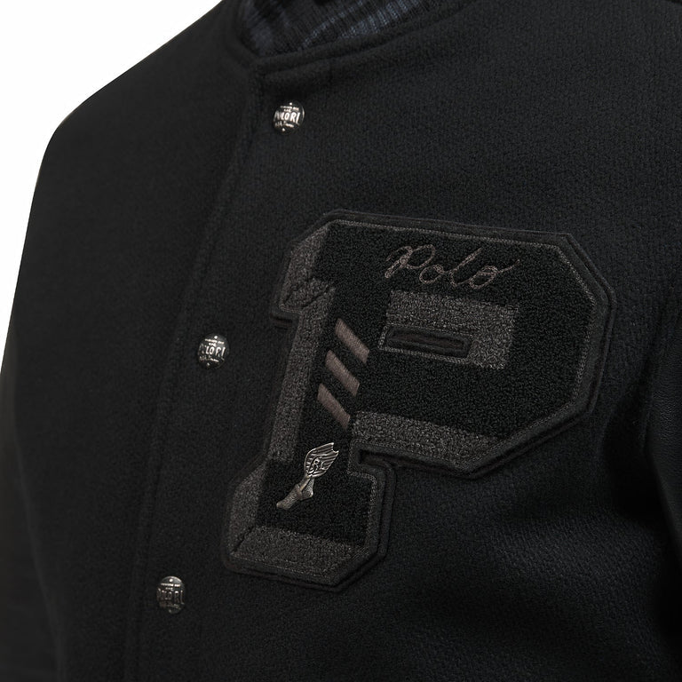 A close-up of the chest area of the black Polo Ralph Lauren Iconic Letterman Jacket. The image showcases the large, black chenille "P" varsity patch with grey "Polo" script embroidery and a small silver-toned winged foot emblem. Detailed view of the silver snap buttons embossed with "Polo RL" and the melton wool texture of the jacket body.